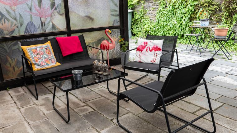 Ein gemütlicher Sitzbereich im Freien mit schwarzen Gartenmöbeln und bunten Kissen. Umgeben von Pflanzen und einer charmanten Steinterrasse.