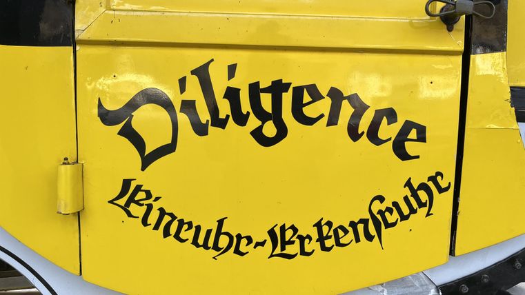 Ein gelbes Fahrzeug mit der Aufschrift "Diligence". Die Schrift ist in schwarzer, verschnörkelter Schriftart gestaltet.