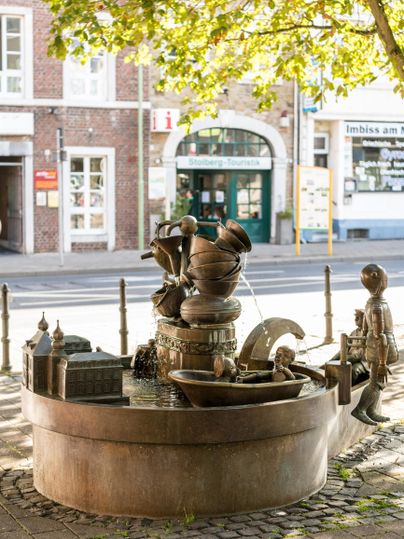 Ein kunstvoller Brunnen mit Figuren, die aus verschiedenen Schalen Wasser schöpfen. Umgeben von Bäumen und nahe einem Gebäude in der Stadt.