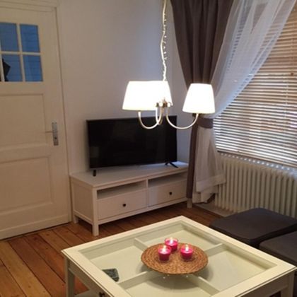 Ein gemütlicher Wohnraum mit einem Fernseher, einem Couchtisch und einer modernen Lampe. Die Fenster sind mit Vorhängen und Jalousien ausgestattet, und der Boden ist aus Holz.