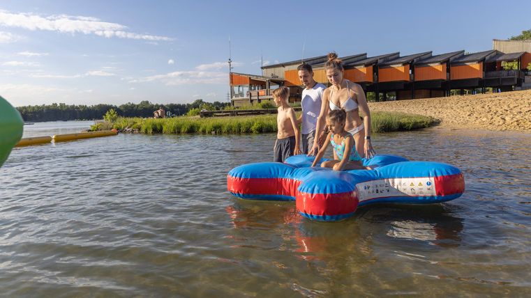 Eine Familie steht auf einer großen aufblasbaren matte im Wasser. Im Hintergrund sind Gebäude und eine ruhige Landschaft zu sehen.