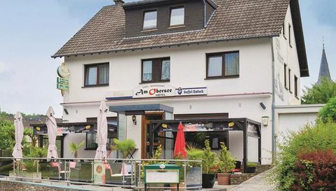 Ein gemütliches Gasthaus mit einer Terrasse, auf der Tische und Sonnenschirme stehen. Im Hintergrund ist ein ruhiger Ort mit grüner Landschaft zu sehen.