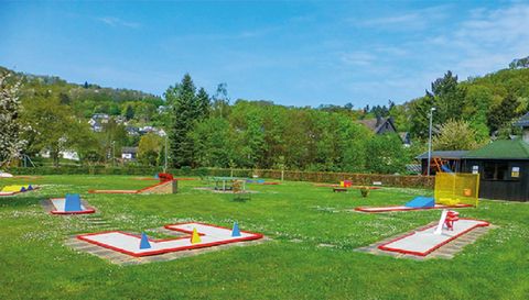 Ein Minigolfplatz in einem grünen Park, umgeben von Bäumen und Hügeln. Die Spieler können auf verschiedenen Bahnanspielen spielen.