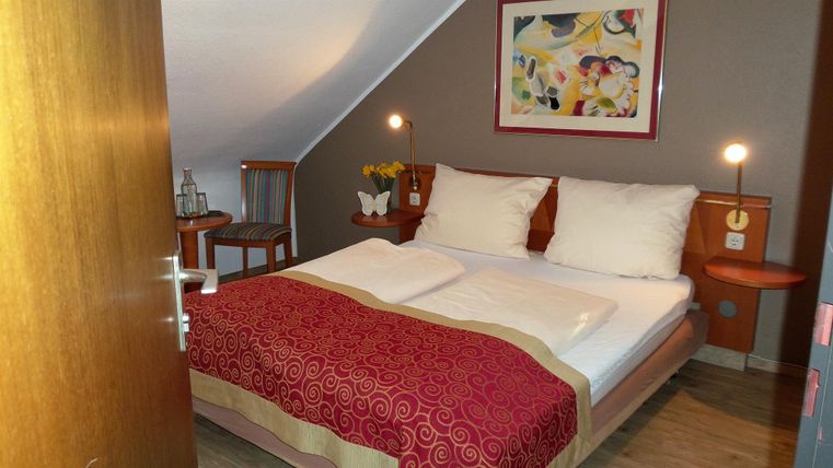 Ein gemütliches Schlafzimmer mit einem Doppelbett und bunten Kissen. An der Wand hängt ein farbenfrohes Bild, und es gibt einen kleinen Tisch mit Blumen.