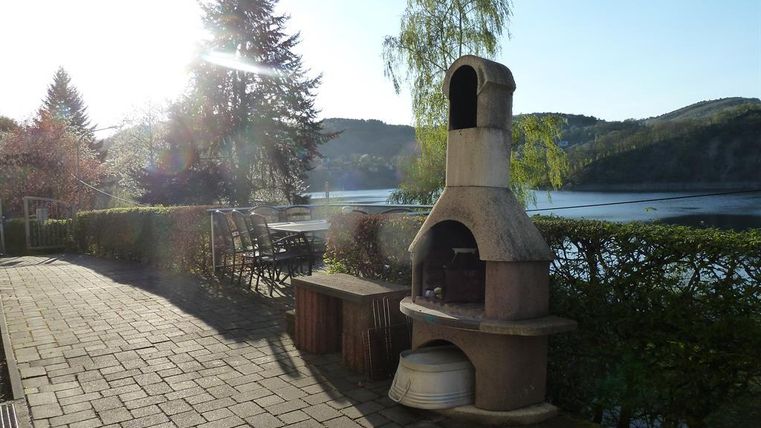 Ein schöner Außenbereich mit einem Grill und Blick auf einen See. Im Hintergrund ist die Natur mit Bäumen und sanften Hügeln zu sehen.