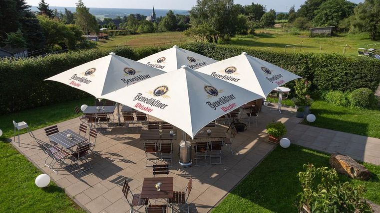 Eine gemütliche Terrasse mit Sonnenschirmen und Holzstühlen. Im Hintergrund ist eine grüne Landschaft zu sehen.