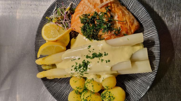 Ein Teller mit gegrilltem Lachs, Spargel und kleinen Kartoffeln. Dazu sind Zitronenscheiben und frische Kräuter dekoriert.