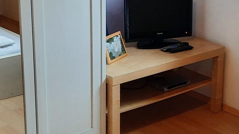 Ein einfacher Raum mit einem Holzschreibtisch und einem schwarzen Fernseher. Neben dem Fernseher steht ein Bilderrahmen auf dem Tisch.