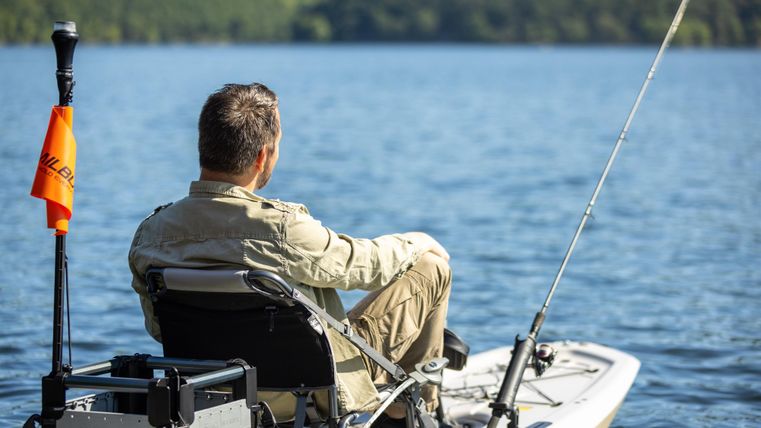 Ein Mann sitzt auf einem Boot und fischt auf einem ruhigen Gewässer. Die Landschaft im Hintergrund ist grün und malerisch.