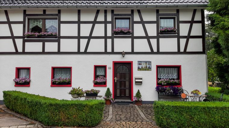 Ein hübsches Fachwerkhaus mit weißer Fassade und roten Fenstern. Vor dem Haus gibt es gepflegte Pflanzen und einen gepflasterten Weg.