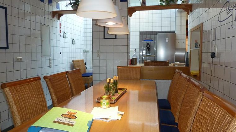 Ein gemütlicher Essbereich mit einem langen Holztisch und Stühlen aus Rattan. Die Wände sind gefliest, und es hängen helle Lampen über dem Tisch.