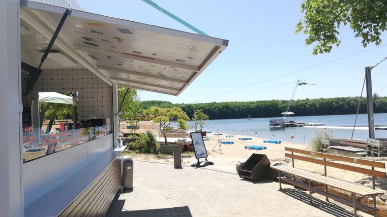 Ein Kiosk am Wasser mit einer schönen Aussicht auf den Strand und den See. Im Hintergrund sind Bäume und Strandliegen sichtbar.
