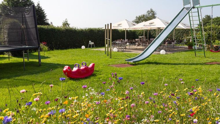 Ein schöner Garten mit bunten Blumen und einer Rutsche. Im Hintergrund sind ein Trampolin und Tische unter Sonnenschirmen zu sehen.